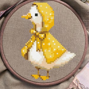May include: Cross-stitch embroidery of a duckling wearing a yellow hooded cape with white polka dots. The duckling has a yellow beak and feet, and is set against a neutral-toned fabric background, framed in a wooden embroidery hoop.