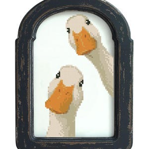 May include: A framed cross-stitch artwork featuring two white geese with orange beaks. The geese are set against a white background within a distressed black frame. The artwork has a rustic, handmade aesthetic.