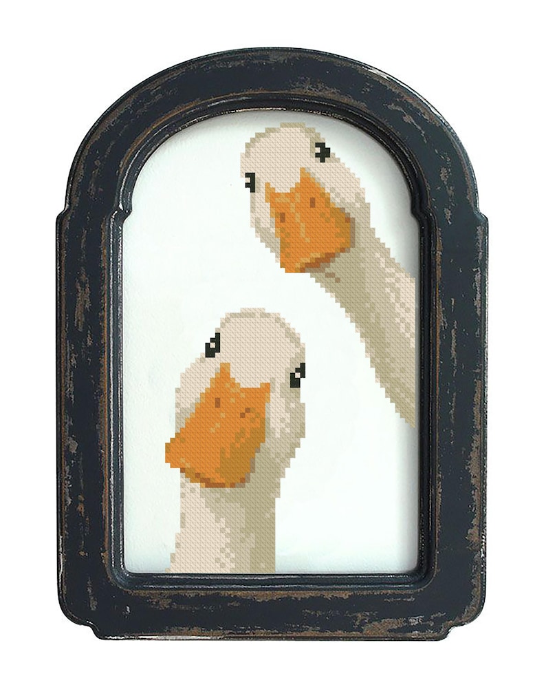 May include: A framed cross-stitch artwork featuring two white geese with orange beaks. The geese are set against a white background within a distressed black frame. The artwork has a rustic, handmade aesthetic.