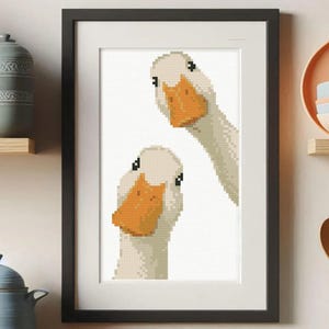 May include: Framed art print featuring a pixelated illustration of two white geese with orange beaks and black eyes. The artwork is set against a white background and framed in a black frame. The print is a whimsical addition to any home decor.