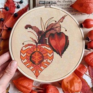 Könnte beinhalten: Ein Kreuzstich-Stickrahmen mit einem detaillierten Design einer stilisierten orangefarbenen und braunen Pflanze. Das Design ist auf einem beigefarbenen Stoff angebracht, mit einem Holzreifen. Das Bild ist von Herbstlaub und Beeren umgeben.