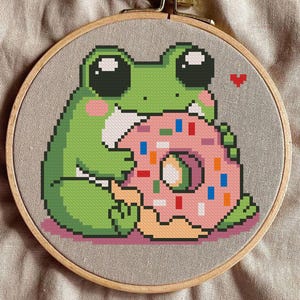 May include: Cross-stitch embroidery of a green frog eating a pink donut with colourful sprinkles. The frog has large black eyes and pink cheeks. The design is set in a wooden embroidery hoop.