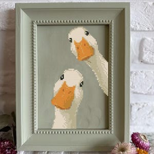 May include: Cross-stitch artwork of two white geese with orange beaks, framed in a light green, beaded-edge frame. The background is a muted green. The artwork is displayed against a white brick wall, with dried flowers at the bottom.