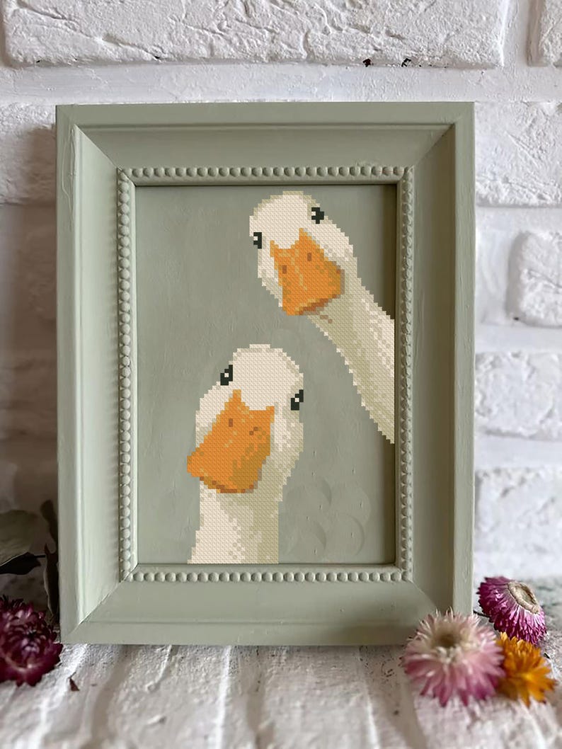 May include: Cross-stitch artwork of two white geese with orange beaks, framed in a light green, beaded-edge frame. The background is a muted green. The artwork is displayed against a white brick wall, with dried flowers at the bottom.