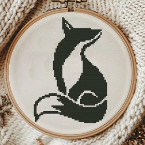 May include: A cross-stitch embroidery of a fox in a wooden hoop. The fox is dark green against a cream-coloured background. The hoop is displayed on a cream-coloured knitted fabric.