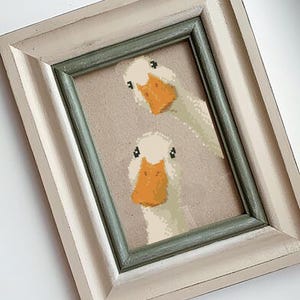 May include: Framed cross-stitch art featuring two white geese with orange beaks and black eyes. The artwork is set against a neutral background and framed in a distressed white and green frame. A charming piece of home decor.