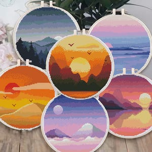 Puede incluir: Cinco aros de bordado redondos con diferentes escenas de puesta de sol. Cada aro presenta un paisaje colorido con montañas, un sol y nubes. Los aros son todos blancos con un marco de madera.