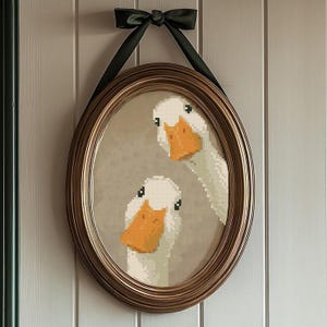 May include: An oval-shaped framed artwork featuring a cross-stitch design of two white geese with orange beaks. The frame is a warm gold color, and the artwork is suspended by a dark green ribbon tied in a bow at the top.