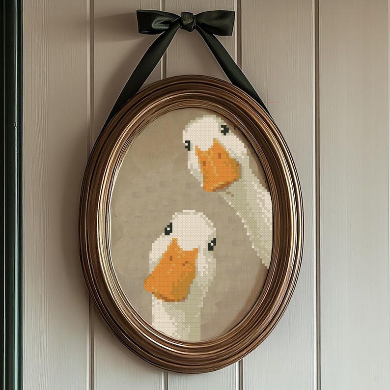 May include: An oval-shaped framed artwork featuring a cross-stitch design of two white geese with orange beaks. The frame is a warm gold color, and the artwork is suspended by a dark green ribbon tied in a bow at the top.