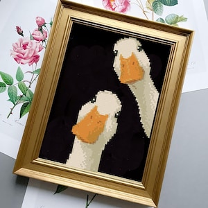 May include: A framed artwork featuring a pixelated image of two white geese with orange beaks against a black background. The artwork is in a gold-colored frame. Floral prints are visible in the background.