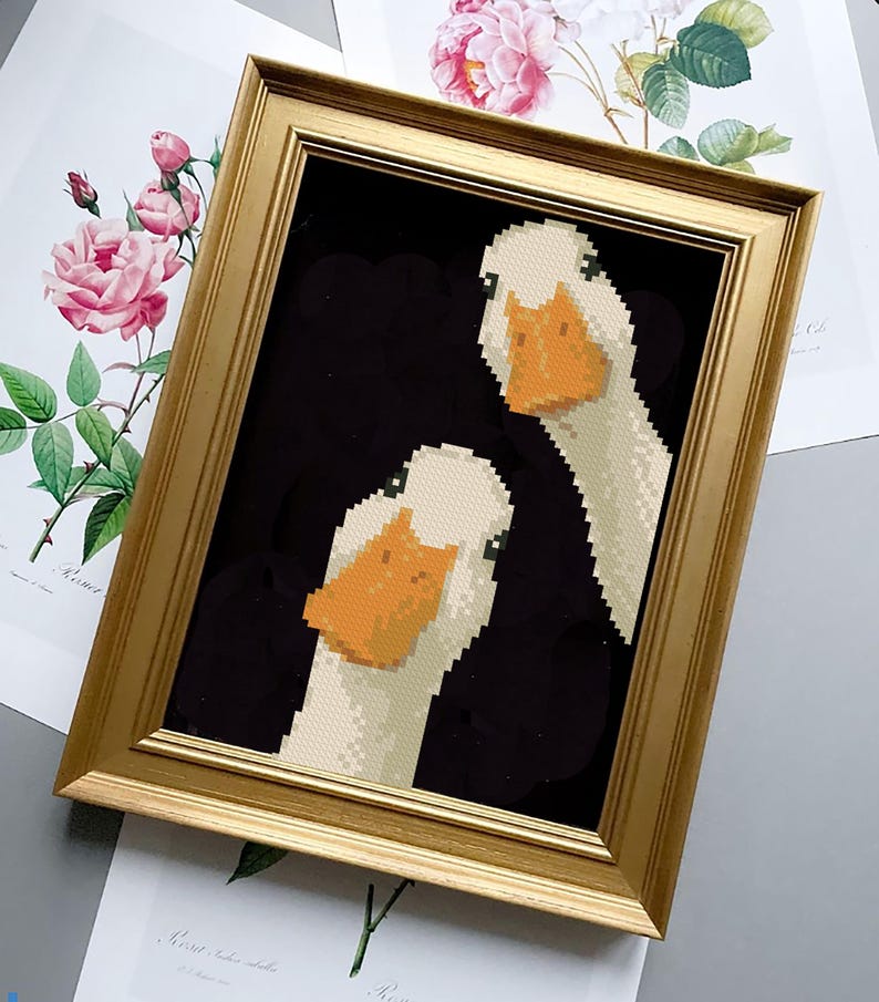 May include: A framed artwork featuring a pixelated image of two white geese with orange beaks against a black background. The artwork is in a gold-colored frame. Floral prints are visible in the background.