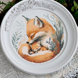 May include: A cross-stitch embroidery depicting a mother fox and her cub curled up together, sleeping. The fox is orange and white, with a bushy tail. The cub is smaller and has a white chest. The background is a light blue with green leaves.