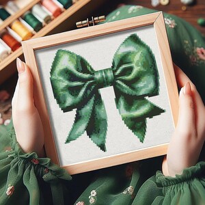 May include: A green bow with a white background in a wooden frame. The bow is made of fabric and has a satin finish.
