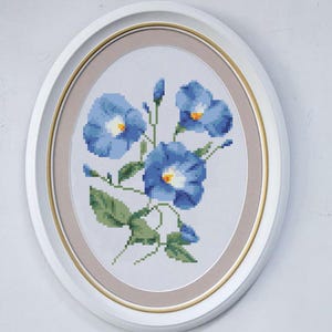 May include: Oval framed cross-stitch art featuring blue flowers with yellow centers and green leaves. The artwork is set against a white background, framed with a white and gold oval frame. A floral themed home decor item.