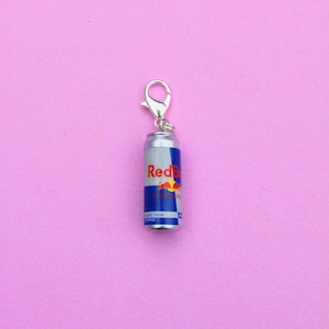 May include: A miniature Red Bull can charm with a silver clasp. The can is silver with blue and red branding, including the word "Red Bull" and a red bull logo. The charm is on a pink background.
