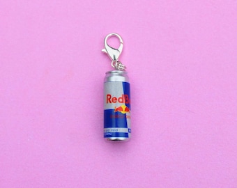 May include: A miniature Red Bull can charm with a silver clasp. The can is silver with blue and red branding, including the word "Red Bull" and a red bull logo. The charm is on a pink background.