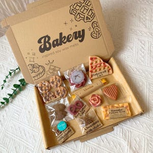 May include: A cardboard box labeled Bakery inspired soy wax melts containing various food-shaped wax melts in clear plastic bags. Items include waffle shapes, a Bakewell Tart, a mocha bar, a heart, a pretzel, and a Banana Pancake Stack.