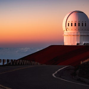 May include: A white observatory dome sits atop a red volcanic hill at sunset. A winding road leads up to the observatory, with a fence lining the side of the road.