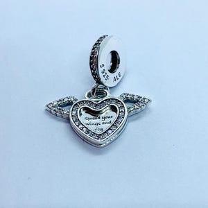 May include: A silver charm with a heart-shaped pendant and wings. The pendant has the text "Spread your wings and fly" and is encrusted with small, clear stones. The charm has a ring for attachment and is marked with "S925 ALE".