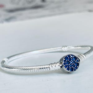 May include: A silver snake chain bracelet featuring a circular clasp centerpiece. The clasp is decorated with a cluster of small, round, deep blue stones set against a metallic silver background, creating a textured and vibrant focal point.