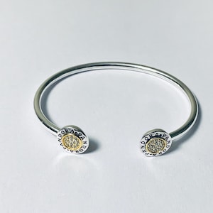 May include: A silver open cuff bracelet featuring two circular end caps. Each cap has a gold-toned center inlaid with small clear stones, surrounded by a silver border engraved with the word PANDORA repeated around the entire circumference.
