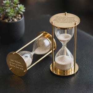 Puede incluir: Dos relojes de arena de metal dorado con bulbos de vidrio transparente llenos de arena blanca. Uno está de pie, grabado con deseos de jubilación. El otro está acostado, grabado con Jane Williams, 35 Years of Service, Retired June 2026.