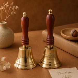 May include: Two hand bells with polished brass bodies and dark brown wooden handles. The right bell features an engraving that reads: Dad, Ring once for coffee and Twice For Hugs, Love Janet. The bells have a flared base and a rounded top finial.