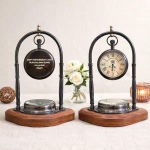 May include: Two arched metal stands with wooden bases, each holding a circular object. One is a clock with a white face and Roman numerals labeled Victoria London 1876. The other is a dark circular plaque engraved with Happy 70th Birthday Linda.