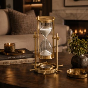 May include: A brass hourglass with white sand, featuring a rotating frame mounted on a circular base shaped like a ship's wheel. The hourglass is positioned on a dark wooden surface, with a compass integrated into the center of the base.