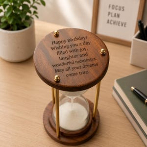 May include: A wooden hourglass with gold-toned metal supports and a clear glass bulb containing white sand. The top circular wooden piece is engraved with the text: Happy Birthday! Wishing you a day filled with joy, laughter and wonderful memories. May all your dreams come true.