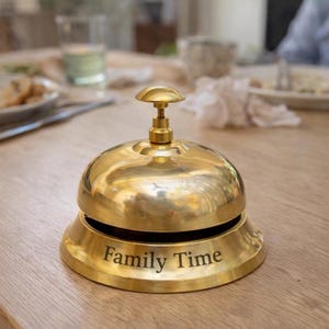 Puede incluir: Un timbre de escritorio de latón pulido con una cúpula redondeada y un pequeño botón en la parte superior. La base del timbre presenta el texto grabado Family Time en una fuente clara y oscura. La superficie metálica tiene un acabado brillante.