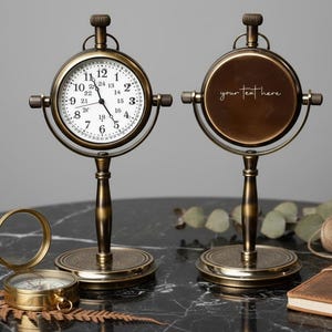 May include: Vintage brass desk clock with a white clock face and black numerals. The clock has a round face and is supported by a decorative stand. The second clock has a blank face with the text "your text here". A compass is also visible.