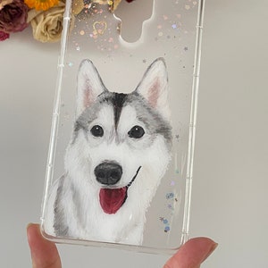 Könnte beinhalten: Transparente Handyhülle mit handgemaltem sibirischen Husky-Design. Die Hülle hat Glitzer-, Stern- und Herzdekorationen. Der Hund hat graues und weißes Fell, schwarze Augen und eine rote Zunge. Die Hülle ist für ein Smartphone.