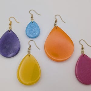 May include: Five teardrop-shaped earrings in shades of purple, blue, orange, yellow, and pink. Each earring has a gold-colored hook. The earrings are arranged on a white background.
