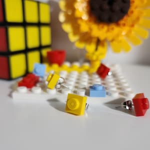 May include: A set of colorful Lego earrings, including yellow, blue, and red pieces. The earrings are displayed on a white base with a white and yellow Lego baseplate.