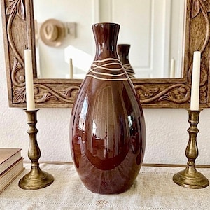 May include: A glossy, dark brown ceramic vase with a narrow neck and a rounded body. The upper portion of the vase features a decorative pattern of thin, light-colored horizontal lines wrapped around the neck area, creating a subtle contrast.