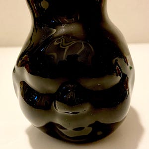 May include: A small, glossy black ceramic vase with a rounded body and a narrow neck. The vase has a smooth, reflective surface, with subtle variations in the black color. The design is simple and elegant, suitable for displaying small flowers or as a decorative item.