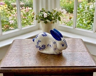 May include: A white ceramic rabbit figurine featuring blue painted floral patterns on its body and solid blue ears. The rabbit is depicted in a seated, resting position with a smooth, glossy finish and detailed blue eyes and facial markings.