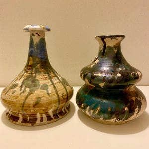 May include: Two ceramic vases with unique shapes and hand-painted designs. The vase on the left has a bulbous base and a long neck, with brown, blue, and green drips. The vase on the right is a double-lobed shape with teal and brown accents.