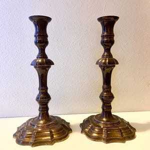 May include: Two antique bronze candlesticks. Each candlestick has a detailed, ornate design with a wide, scalloped base and a slender, tiered stem. The candlesticks are a warm, dark bronze color, adding a touch of vintage elegance.