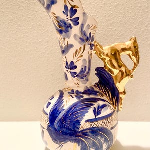 May include: A white ceramic pitcher with blue floral and rooster designs, accented with gold trim. The handle is a gold-colored animal figure. The pitcher has a unique, curved spout.