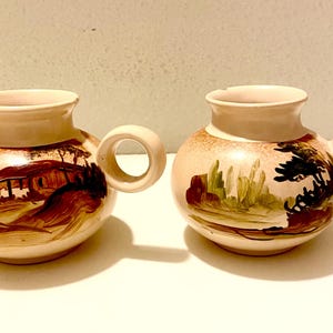 May include: Two ceramic mugs with round handles. The mugs are beige with hand-painted landscape scenes in brown, green, and black. The mugs have a bulbous shape and a wide opening.
