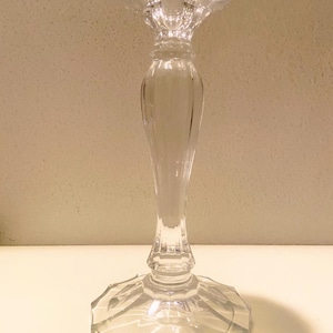May include: A clear glass candlestick holder with a faceted base and a decorative, fluted stem. The top of the candlestick has a flared, rounded shape. The candlestick is designed to hold a single candle and is suitable for home decor.