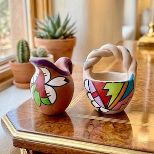 May include: A small terracotta pitcher with a purple rim and a painted white flower design alongside a small, round, light-colored clay basket featuring a braided handle and a vibrant, multi-colored geometric pattern painted on its exterior surface.