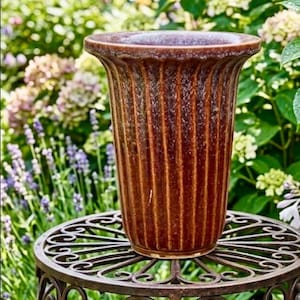 May include: A brown, ribbed ceramic planter sits on a decorative wrought iron plant stand. The planter has a wide rim and a textured surface. The background features lush greenery and flowers, creating a garden setting.