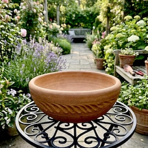 May include: A round terracotta bowl with a thick, rolled rim and a decorative band of carved, slanted grooves encircling the exterior. The bowl is placed on a black metal stand featuring intricate, swirling scrollwork patterns.