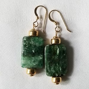 以下が含まれることがあります： 金色のアクセントが付いた緑色の石のイヤリングのペア。イヤリングは長方形で、表面にテクスチャがあります。