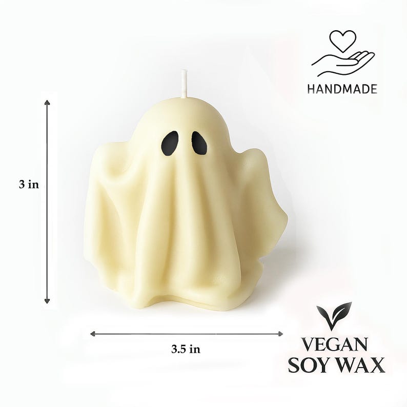 May include: A handmade, ivory-colored ghost-shaped candle, 3 inches tall and 3.5 inches wide, with black oval eyes. The candle is made from vegan soy wax. The image includes the text "HANDMADE" and "VEGAN SOY WAX".