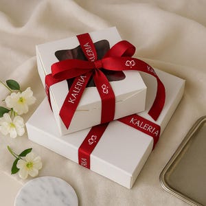 May include: Two white gift boxes are stacked, one atop the other. The top box has a clear window, revealing a dark interior. Both boxes are adorned with a red ribbon printed with "KALERIA" and a crown symbol. The ribbon is tied in a bow on the top box.
