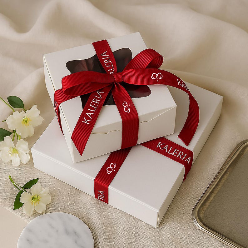 May include: Two white gift boxes are stacked, one atop the other. The top box has a clear window, revealing a dark interior. Both boxes are adorned with a red ribbon printed with "KALERIA" and a crown symbol. The ribbon is tied in a bow on the top box.